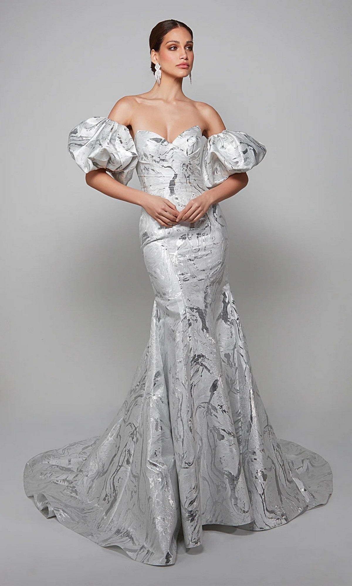 A woman poses against a plain grey background in the Alyce 7055—a long white dress featuring dramatic puff sleeves and a fitted, floor-length skirt, ideal for mothers-of-the-bride seeking timeless elegance.